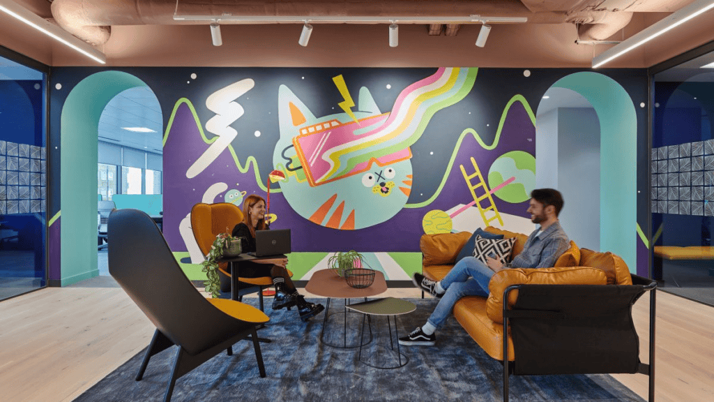 The image shows a sensory-friendly break room for office workers. The room is well-lit and has various tints and hues of blue elements in it. The wall in the front has an illustration of a cat and mountains and other things, while the walls on either side seem to be made of glass. The room has a wooden veneer floor with a dark blue rug in the middle. On the rug are 2 chairs and a sofa, along with 2 small coffee tables. There are 2 plants to provide some greenery to the room. There is a man seated on the sofa, with his legs crossed and he is smiling at a woman seated on the chair which is closer to the wall. She is smiling at him while working on her laptop. There are 2 arch entrances on either side of the wall up front. 