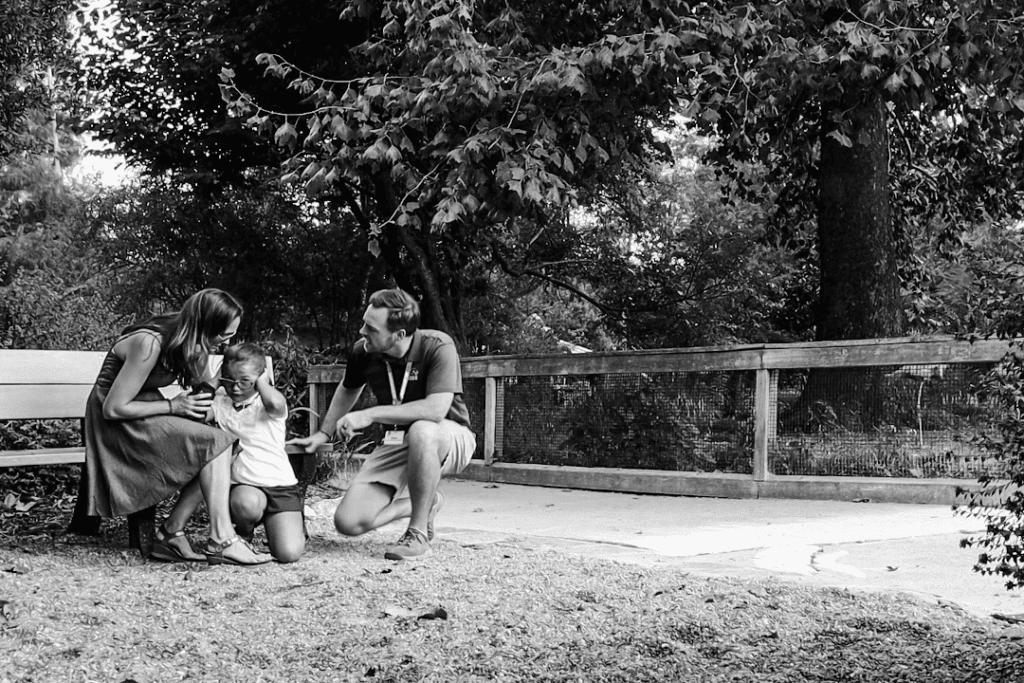 The black-and-white image shows a young neurodivergent child crouching, probably dealing with sensory overload, between two adults who appear to be comforting the child. The adult woman on the left is gently leaning in and patting the child’s back, She is seated on a bench while the child is crouched down holding their head in their hands, covering their ears. The child is leaning towards the woman, while the adult man on the right offers support with a hand extended. He is crouching beside the child and offering his support. The trees and a wooden fence create a natural, outdoor background. The adults are presumably the child’s parents.