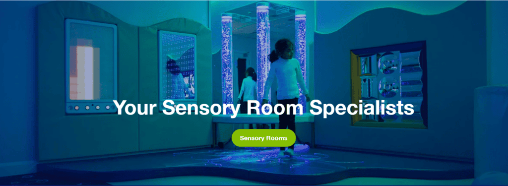 A sensory room illuminated with a calming turquoise-blue light features interactive panels and central bubble tubes. The smooth blue floor and curved walls create a soothing, immersive atmosphere, with the phrase "Your Sensory Room Specialists" prominently displayed below.
