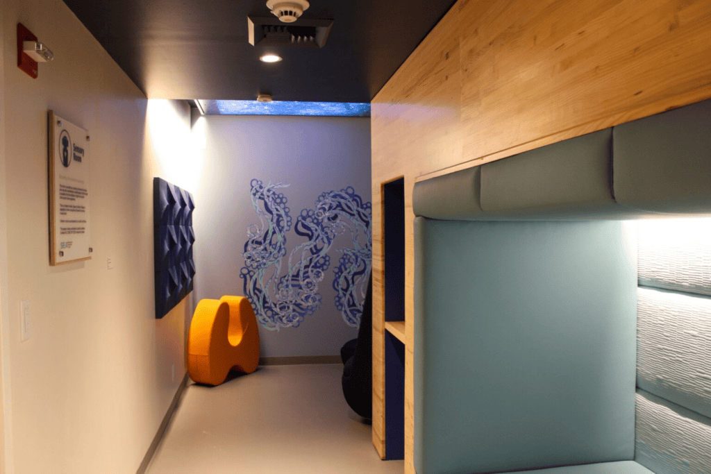 The image shows a calming sensory room set up in the Seattle-Tacoma International Airport, with calm lighting, interesting tactile textures and temporary art walls, with soft, padded seating and acoustic panels on the walls. There is a brightly coloured, abstract orange chair and blue wall art resembling organic shapes, providing a calming environment. The ceiling has a textured light source, contributing to the sensory experience. A sign indicates the room's purpose.