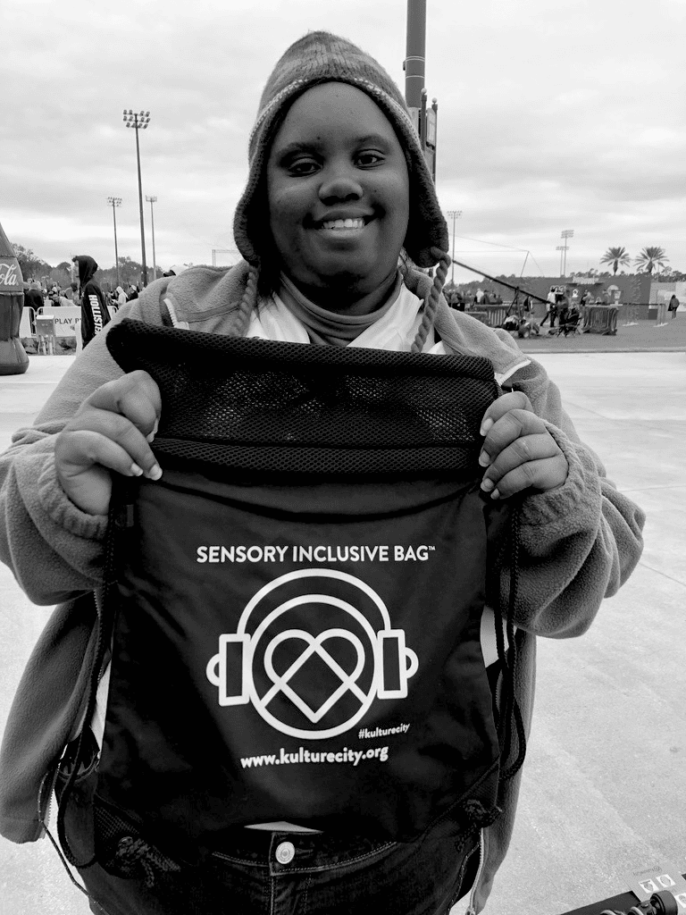 A person stands outdoors, smiling warmly at the camera, holding up a black bag with "Sensory Inclusive Bag" written on it. The bag features the Kulture City logo—a pair of headphones with a heart inside. The background shows a stadium and cloudy skies.