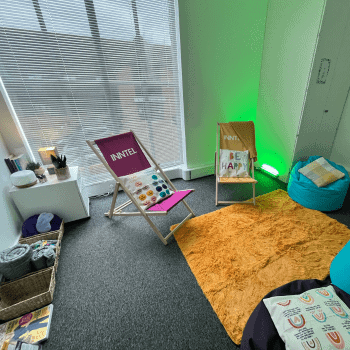 The image shows a sensory quiet room in a workplace environment. The room is well-lit with baskets containing towels and other sensory tools. The floor has a textured rug and two foldable chairs with a cushion on top. There are a few small teal bean bags with cushions on them. The room has white walls and a grey textured mat covering the floor. The overall feel is calming and peaceful.