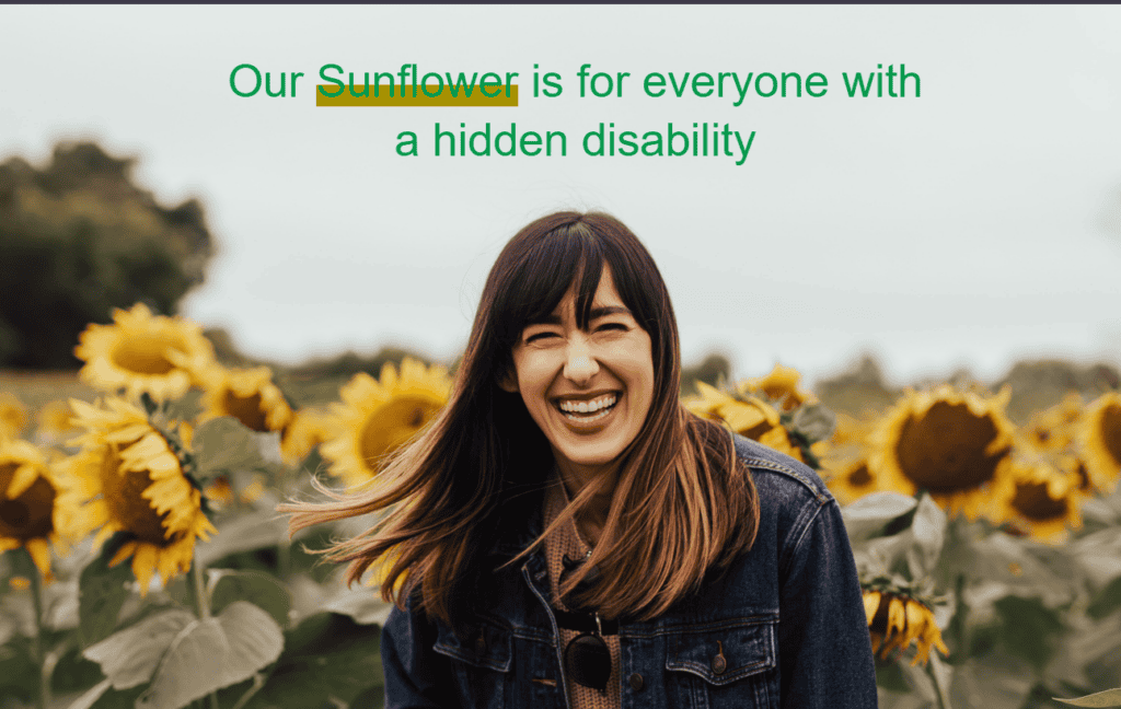 A woman is smiling brightly while looking towards the camera, in the midst of a sunflower field. In the top middle, where the sky is visible in a blurred background, it says “ Our Sunflower is for everyone with a hidden disability” with a mustard-colored thick underline under the word “Sunflower”.