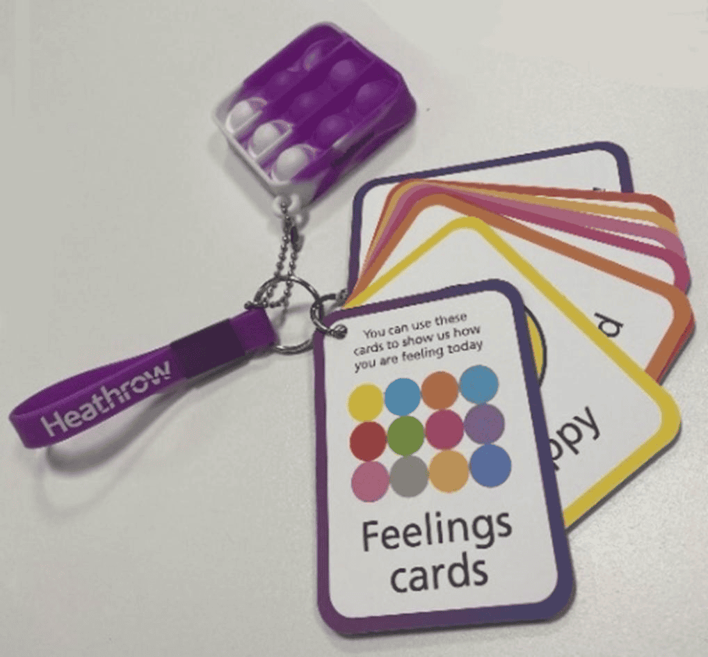 The image shows a sensory area at London Heathrow Airport. On the left, a woman is sitting in a padded space with colourful, cityscape-themed wall art and a mirror. On the right, a keychain holds "Feelings cards" designed to help individuals with hidden disabilities express their emotions.
