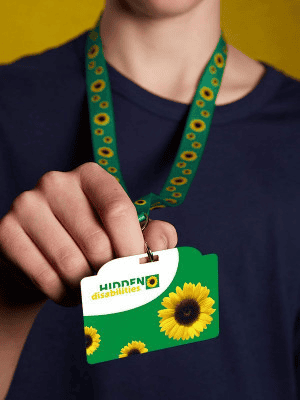 The image shows a person wearing a lanyard around their neck with a Hidden Disabilities card attached. The person, dressed in a black T-shirt, is holding the card in their hand, but their face is not visible. The card is divided into two sections: a white upper portion with the words "HIDDEN disabilities" in the top left corner, and a green lower section featuring three sunflowers. This card is part of the services offered by the Hidden Disabilities Sunflower initiative, which discreetly indicates that the wearer may need additional support or understanding.